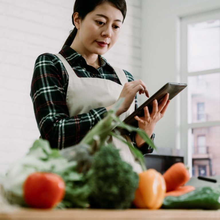 Femme portant un tablier japonais en cuisine consultante une tablette, entourée de légumes frais.