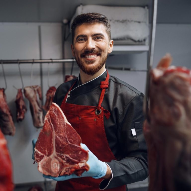 Boucher souriant tenant un morceau de viande, entouré de viandes accrochées.