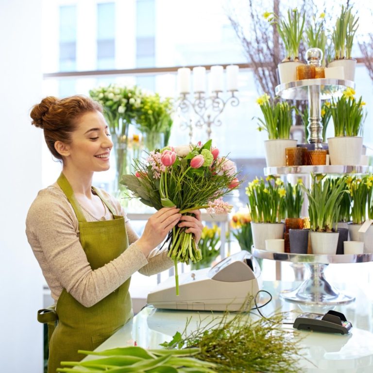 Femme souriante préparant un bouquet de fleurs avec un tablier vert clair dans une boutique lumineuse.