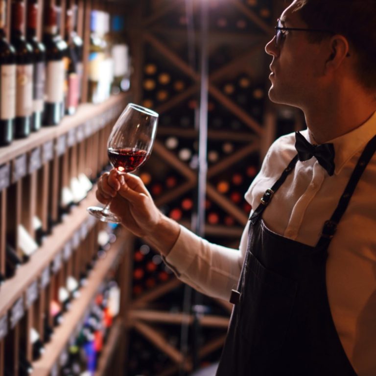Un sommelier examine un verre de vin rouge devant une étagère à vins.